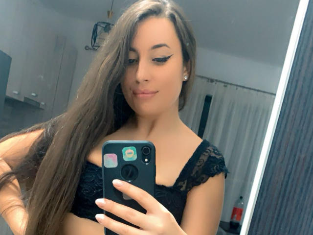 CellineStarX with long brown hair, black lace bra, and winged eyeliner, taking a mirror selfie, showing her tits, holding a black phone, in a bathroom.