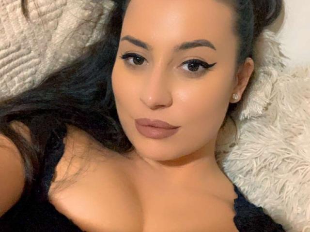 CellineStarX with medium brown skin, dark hair, and thick eyebrows, wearing black lace, has full boobs, and is lying on a fluffy pillow, with sultry eyes and brown lipstick.