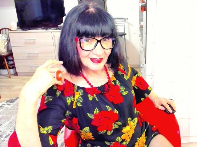 TiniaMartin with black bob, glasses, red lipstick, floral black dress, red bead necklace, red chair, hand holding orange object, smiling, white background, wooden furniture, bright lighting.