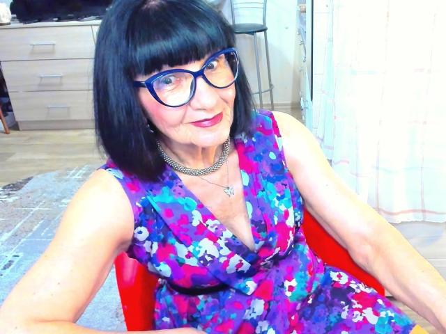 TiniaMartin with black bob, glasses, purple floral dress, red chair, necklace, smiling, middle-aged, pink lipstick, sitting, arms on red chair, light skin, medium-sized tits, inside room.