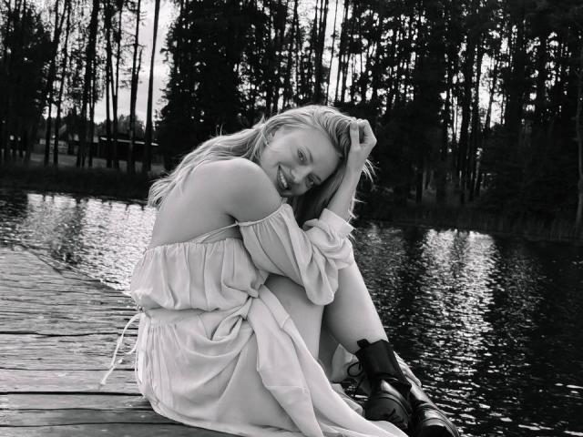 Black-and-white photo of CrystalHope with long blonde hair, smiling, sitting on a wooden dock, wearing an off-shoulder dress and boots, her hand in her hair, by a lake.