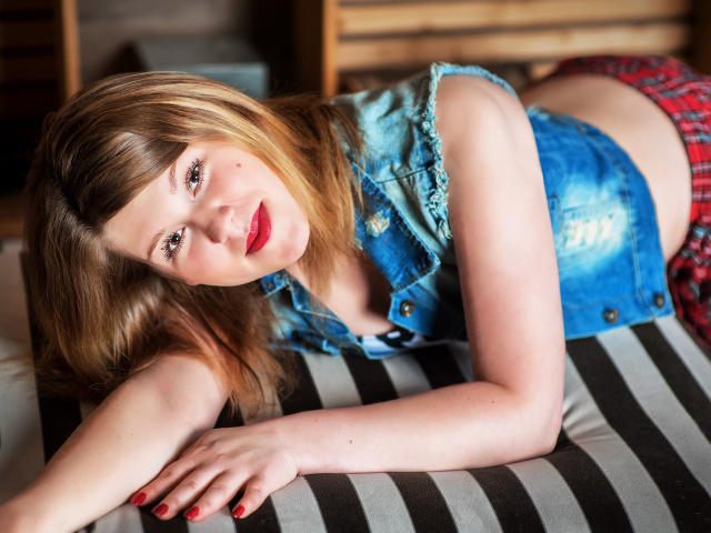 KarolynGlam with blonde hair, blue denim vest, red plaid skirt, lying on a black and white striped pillow, looking at the camera, red lipstick, red nails, smiling.