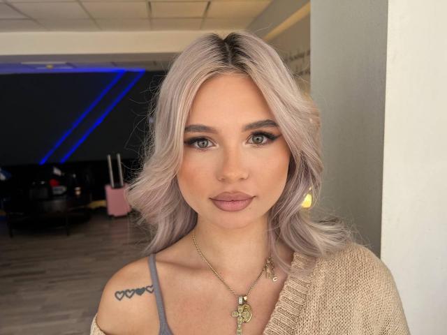 JessyyBelle with platinum blonde hair, green eyes, and pink lips, wearing a gold necklace, showing a tattoo on her left shoulder, and a beige off-shoulder top.