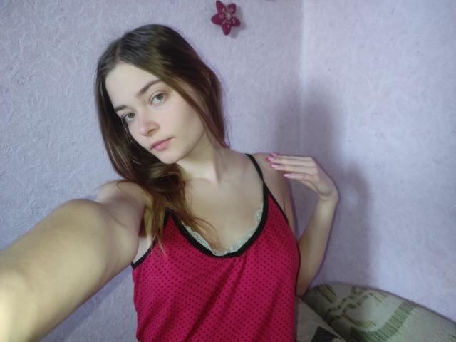 HappyEmily with light skin and brown hair, wearing a pink polka-dot tank top, showing small tits, and pink nails, taking a selfie, hand near collar, looking at camera.