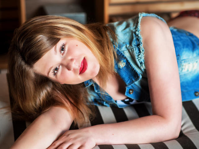 KarolynGlam is lying on a striped black-and-white cushion, her head tilted to the left. She has fair skin, light brown hair with blonde highlights, and red lipstick. She wears a blue, distressed denim sleeveless vest. Her right arm is stretched out in front of her, while her left arm is slightly bent. The lighting is soft, emphasizing her facial features and the texture of her vest. Her expression is relaxed and slightly curious. The composition focuses on her upper body and face. The image has a realistic, photographic style.