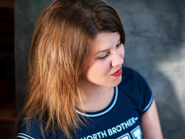 KarolynGlam with shoulder-length blonde hair, red lipstick, and blue shirt, looking sideways with a slight smile. Her ass is not visible. Her tits are small and slightly visible.