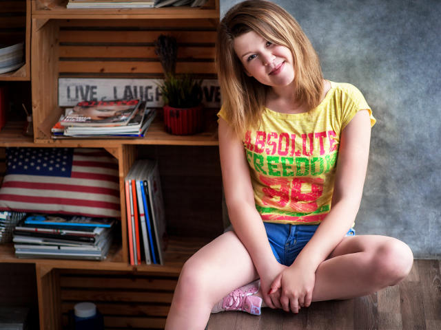 KarolynGlam sits on a wooden table with her legs spread apart. She has light skin, shoulder-length blonde hair, and a smiling expression. She wears a yellow T-shirt with bright pink and green text, and blue shorts. Her hands rest on her pink sneakers. A wooden shelf with books, an American flag, and a "Live Laugh Love" sign is behind her. The image has a watermark in the bottom right corner.