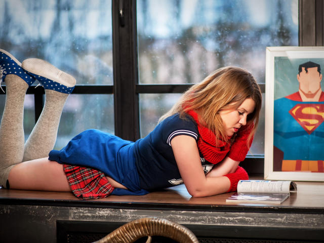 KarolynGlam lies on a wooden table, face down, wearing a blue dress with a red plaid skirt, white socks, and blue polka-dot heels. She has blonde hair, a red knit scarf, and looks at an open book. Her legs are bent at the knees, feet in the air. A framed Superman poster stands next to her. The window behind shows a blurred, snowy outdoor scene. The table has a wicker basket in the foreground. The image has a realistic, yet slightly stylized, photographic quality.