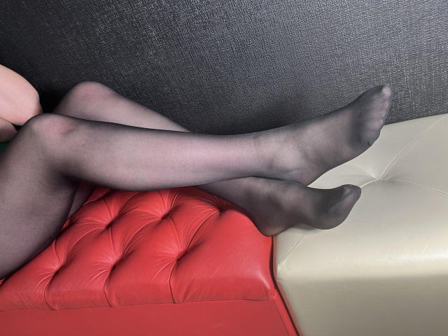 KarolynGlam's legs in sheer black pantyhose, crossed at the ankles, resting on a red and beige tufted leather seat. Her feet are pointed, showing her toes.