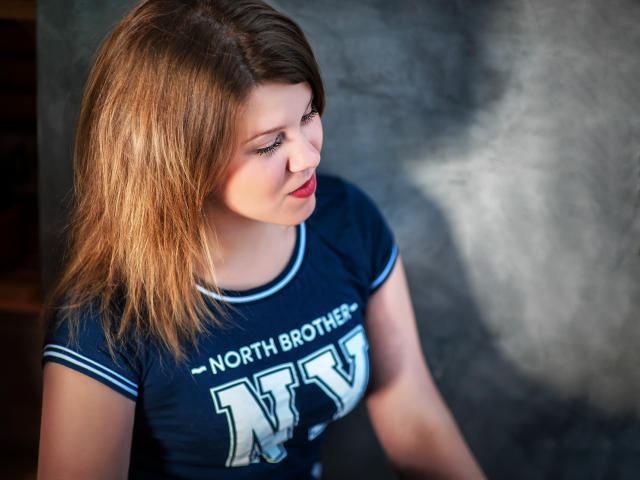 KarolynGlam with blonde hair, blue shirt, red lipstick, looking down, tits slightly visible, soft light, textured wall in background.