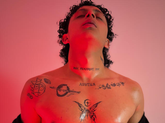 El rostro de Jay Taylor, sin camisa y con el cabello rizado, está inclinado hacia atrás, mostrando tatuajes en su pecho y cuello. La luz roja resalta su torso sudoroso y tatuado, con diversos diseños de tinta..