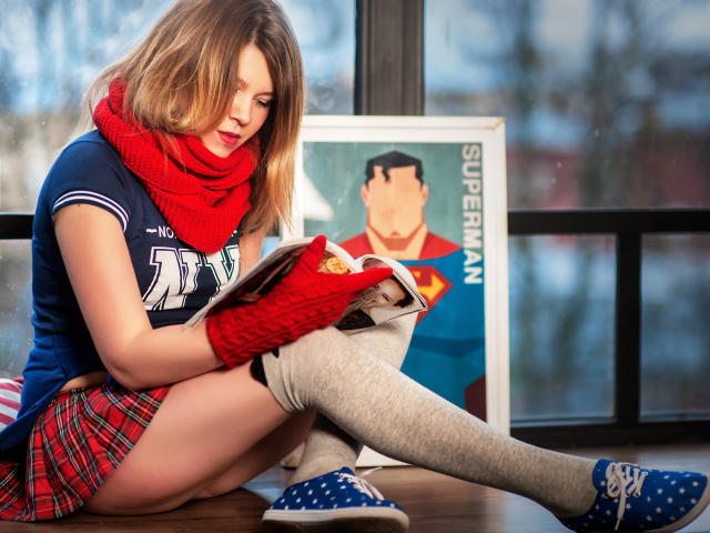 KarolynGlam sits on a wooden floor, reading a comic book. She wears a blue sports jersey, red scarf, red plaid skirt, gray knee socks, and blue sneakers. Her blonde hair is slightly messy. Red gloves cover her hands. A framed Superman poster stands behind her on the right. Natural light from a window illuminates her. The image has a casual, everyday feel.