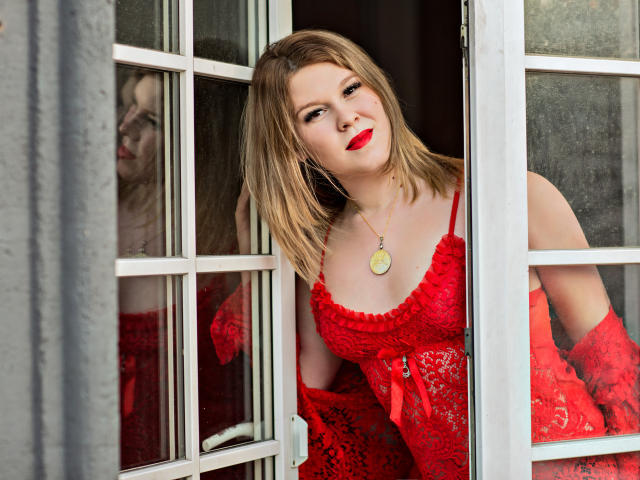 KarolynGlam stands in a partially open window, wearing a red lace dress with a bow at the chest. She has blonde hair, red lipstick, and a confident expression. A gold pendant necklace hangs around her neck. Her right hand holds the window frame while her left hand rests on the glass. The window has white frames and multiple panes. Reflections of KarolynGlam are visible in the glass. The texture of the lace is detailed and intricate. Her red dress contrasts with the gray exterior. The composition focuses on KarolynGlam's upper body and face.