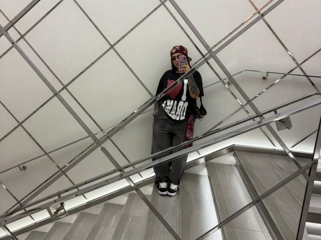 TessaMagic stands on a staircase, wearing a red mask, black shirt with white lettering, gray pants, and white shoes, holding a phone. Metal railings and white walls.