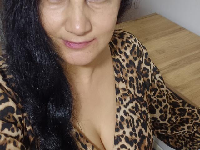 KarinaCecillia with medium brown skin, long black hair, pink lipstick, and leopard print top, showing cleavage, slightly smiling, standing near a wooden surface.
