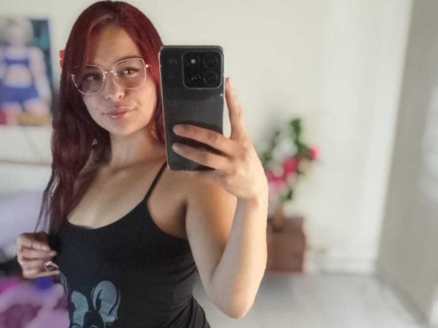 SaraAndMarilyn69 with red hair, glasses, and black tank top showing small tits, taking a selfie with a black phone in a white room.