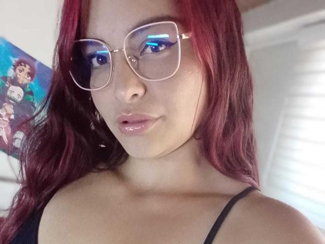 SaraAndMarilyn69 with red hair, glasses, and glossy pink lips, wearing a black spaghetti strap, showing a cute face and blue eye makeup. Boobs partially visible.