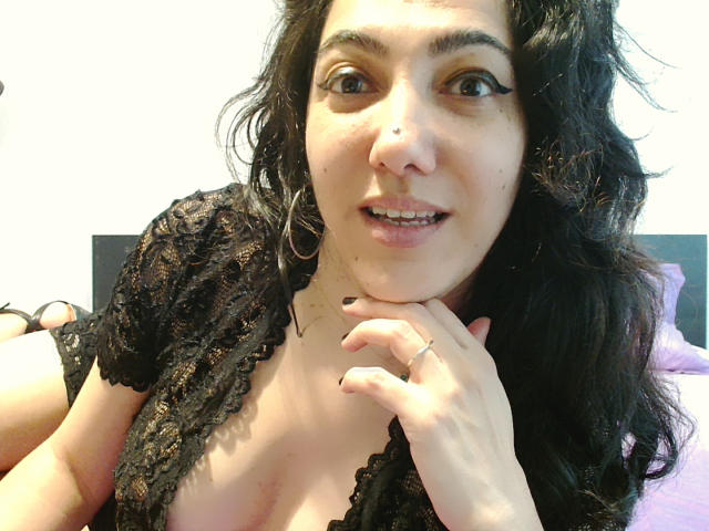 SexyMirra with long, wavy black hair, wearing a black lace top, showing cleavage, black eyeliner, and a ring on her finger, smiling with her hand near her chin.
