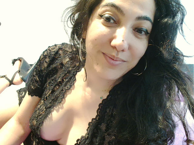 Close-up photo of SexyMirra with wavy black hair, light brown skin, and subtle makeup. She's wearing a black lace top with a deep V-neck, revealing a small portion of her left breast. She has a small nose ring and large hoop earrings. Her brown eyes are slightly upturned, and she's smiling softly. The image has a casual, intimate feel with natural light highlighting her features. Her left arm is visible, holding a black high-heeled shoe. The background is blurred.