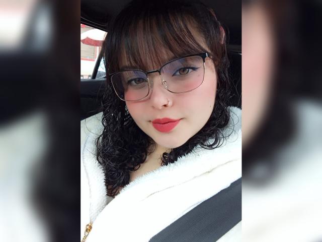 GiaDonovan heeft krullend zwart haar, een bril, rode lippenstift en een witte, pluizige jas, en zit in een auto met een veiligheidsgordel. Ze heeft een neusring aan de linkerkant..