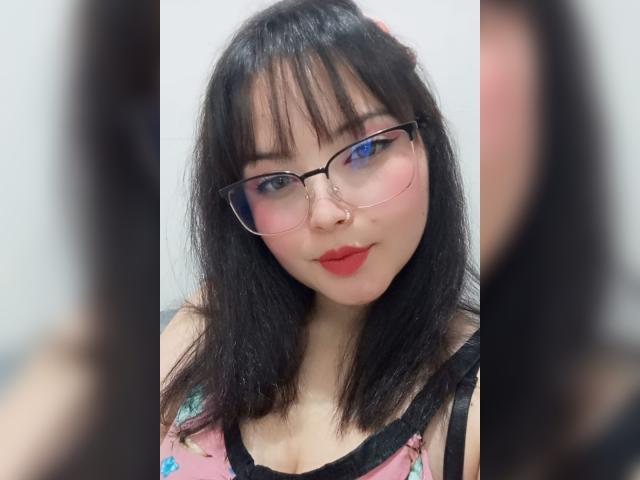 GiaDonovan heeft zwart haar dat tot op haar schouders reikt, draagt een bril, rode lippenstift en een neusring. Ze draagt een roze top met bloemenprint en zwarte randen, waarbij haar borsten zichtbaar zijn. Vage reflecties omlijsten haar..