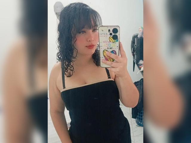 GiaDonovan with curly black hair, red lipstick, black strapless dress, holding phone in mirror selfie, showing medium tits, soft pink skin, and a sexy, confident look.