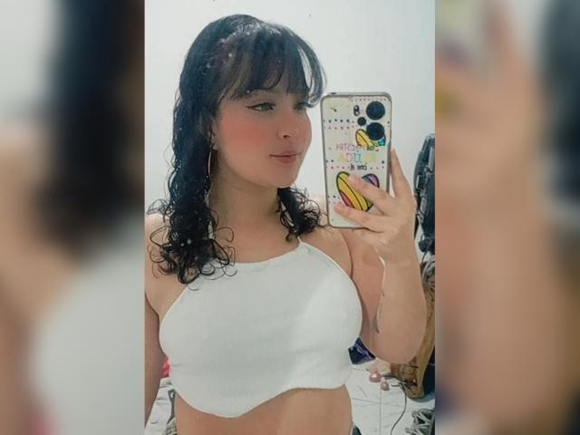 GiaDonovan mit lockigem, schwarzem Haar, trägt ein weißes Top mit Halter und hält ein Handy mit einer bunten Hülle. Sie macht ein Selfie vor einem Spiegel, auf dem ihre Brüste zu sehen sind..