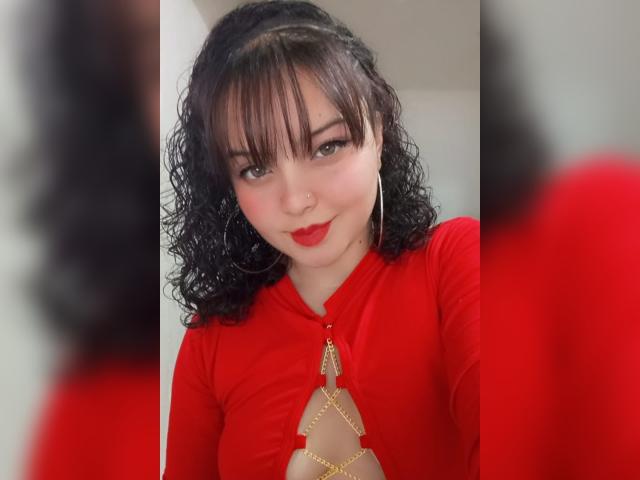 GiaDonovan, com cabelo preto encaracolado, batom vermelho e uma blusa vermelha com um recorte no peito, usando detalhes de corrente dourada e grandes brincos de argola, sorrindo para a câmera..