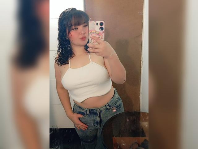 GiaDonovan with curly dark hair, wearing a white spaghetti strap top and blue jeans, holding a phone, showing her belly and hand in her pocket, in a mirror selfie.
