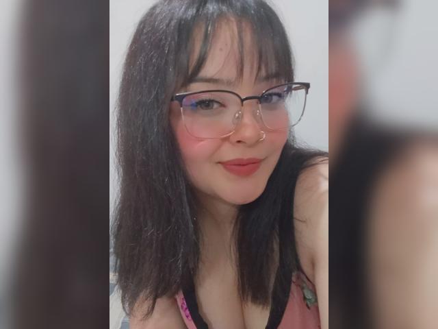 GiaDonovan, con capelli neri, occhiali, piercing al naso e fard rosa, sorride alla fotocamera, mostrando il seno con una maglietta rosa e nera. Sfondo sfocato..