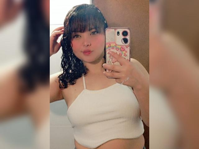GiaDonovan, avec ses cheveux noirs mouillés, ses lèvres roses et pulpeuses, portant un crop top blanc côtelé, tenant une coque de téléphone rose avec des cœurs, reflétée dans un miroir. Seins visibles..