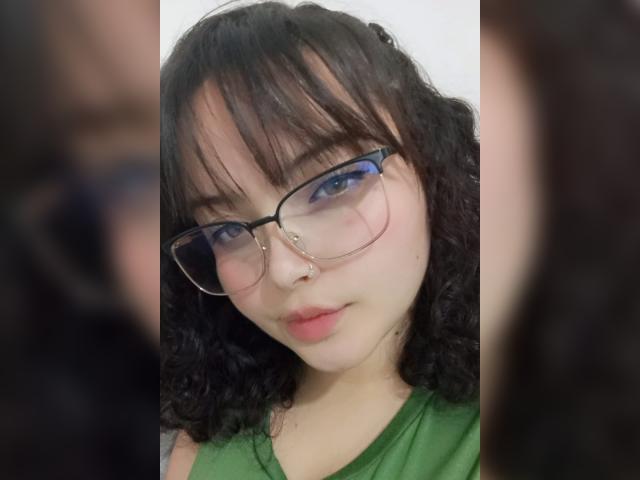 GiaDonovan has curly black hair, blue eyeshadow, and wears glasses with a nose ring. She's wearing a green top, and her pale skin contrasts with her dark hair.