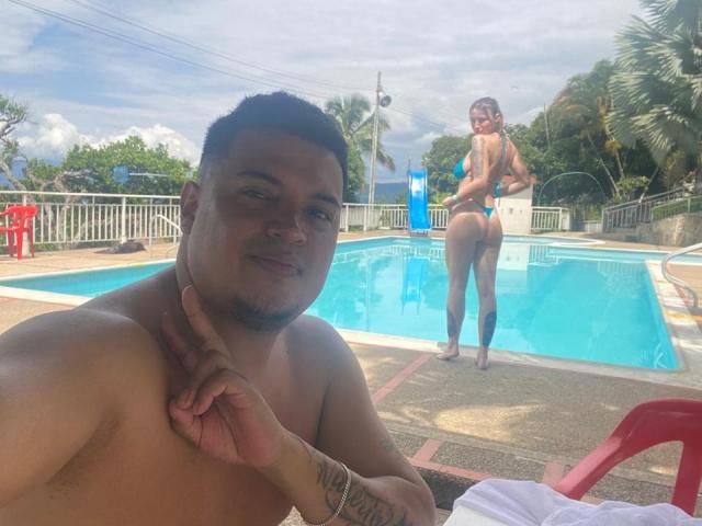 TheManagers with short black hair and a goatee, shirtless, pointing at his neck, tattoo on arm. TheManagers with brown hair, back to camera, showing her ass in a thong.