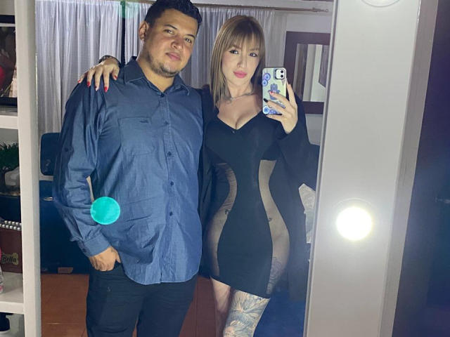 A selfie photograph shows two people standing in front of a mirror. TheManagers, a man with short dark hair in a blue shirt and black pants, stands to the left. He has a relaxed, slightly smiling expression. The second TheManager, a woman with blonde hair and pink lipstick, stands to the right, holding a phone with a colorful case. She wears a black, sheer dress with a deep V-neckline, revealing a tattoo on her right thigh. A bright circular light is visible in the mirror's reflection. The man's hand is on the woman's shoulder.