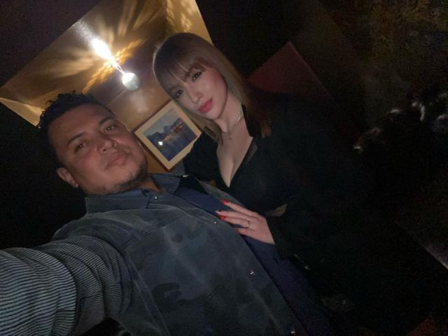 A photograph of TheManagers. A man with short, curly dark hair and a beard stands on the left, wearing a striped shirt and a camo vest. He has a slight smile. A woman with straight, brown hair and bangs stands on the right, wearing a black jacket with a low neckline. She has a hand on TheManagers' chest, with red-painted nails. The woman's expression is neutral. The lighting is dim, with a single overhead light casting shadows. A framed picture is visible in the background.