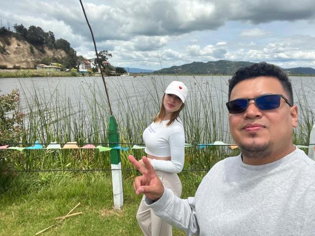 A selfie with two people in a grassy lakeside setting. TheManagers in the foreground has short black hair, wears dark blue sunglasses, and a grey long-sleeve shirt, making a peace sign with his right hand. The second TheManager, in the background, has long brown hair, wears a white cap and white long-sleeve crop top, and beige pants. Colorful flags are strung across a green and white fence. The lake is calm with reeds in the foreground. Overcast sky with mountains in the distance.