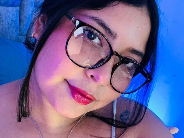 DakotaJhons69 with black glasses, red lipstick, and a slight smile, has a tattoo on her right shoulder, wearing a white strap, and blue lighting.