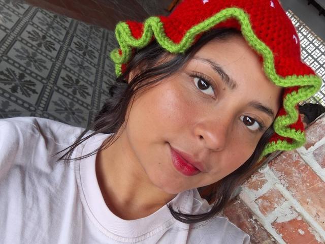 DakotaJhons69 with a red and green crocheted hat, wearing a white shirt, has a cute face, dark eyes, and red lips, standing against a brick wall.