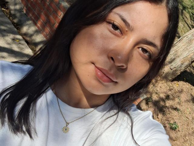 DakotaJhons69 has tan skin, black straight hair, brown eyes, and glossy pink lips. She's wearing a white shirt and a gold necklace with a star-shaped pendant.