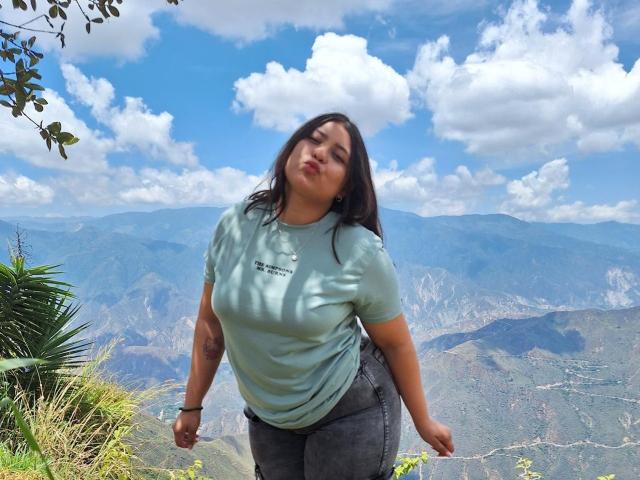 DakotaJhons69, chubby, medium tits, wearing light blue shirt, dark gray pants, pouty lips, black hair, tattoo on left arm, standing against mountain backdrop, blue sky, white clouds.
