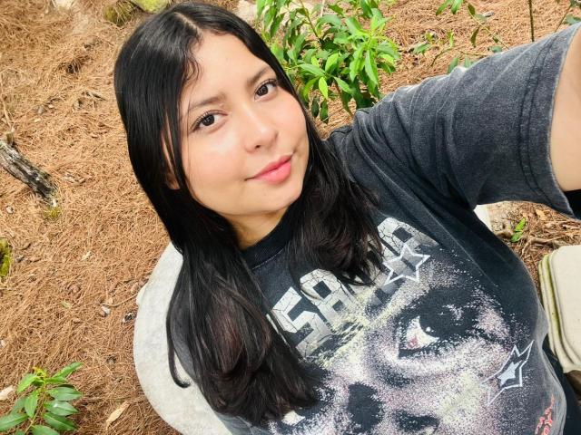 DakotaJhons69, with long black hair, wearing a black "Skid Row" shirt, smiling, has a cute face, and is taking a selfie outdoors, showing a small, subtle smile.