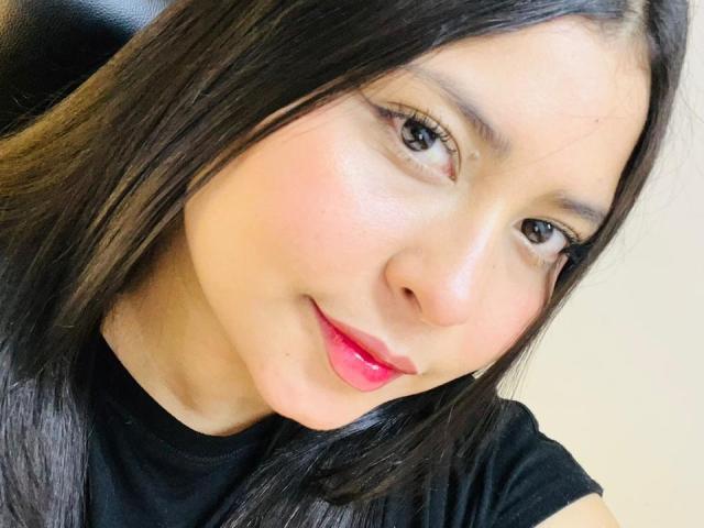 DakotaJhons69 has a cute face with long black hair, pink lips, and a slight smile. She's wearing a black shirt, with fair skin and brown eyes.
