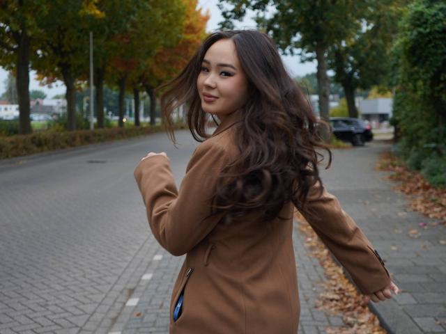 Dbodifka with long brown hair, tan coat, looking back, smiling, standing on a brick-paved street, autumn leaves, overcast day, right arm raised, left arm down.