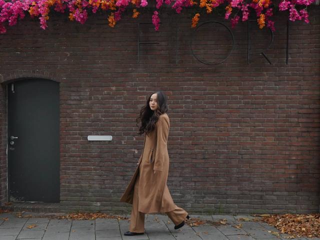 Dbodifka with long brown hair, wearing a brown coat and black shoes, walks against a brick wall with vibrant pink and orange flowers overhead. Her ass is slightly visible.
