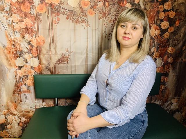 VeronicaSvitt has blonde hair, light skin, and small tits, wearing a blue striped shirt and jeans, sitting on a green chair with hands on her knees.