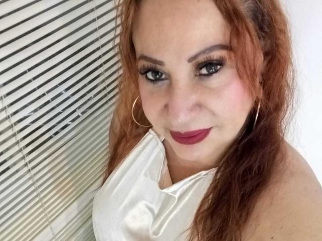 LucianaBlack ha i capelli rossi, orecchini a cerchio e indossa una canottiera bianca, con rossetto rosso scuro, e guarda verso l'alto con un leggero sorriso verso la fotocamera..