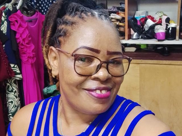Sweetbooty27, con piel oscura, lápiz labial rosa y gafas, vestida con un vestido azul a rayas, tiene el cabello trenzado y una sonrisa segura, mostrando su trasero en el fondo..