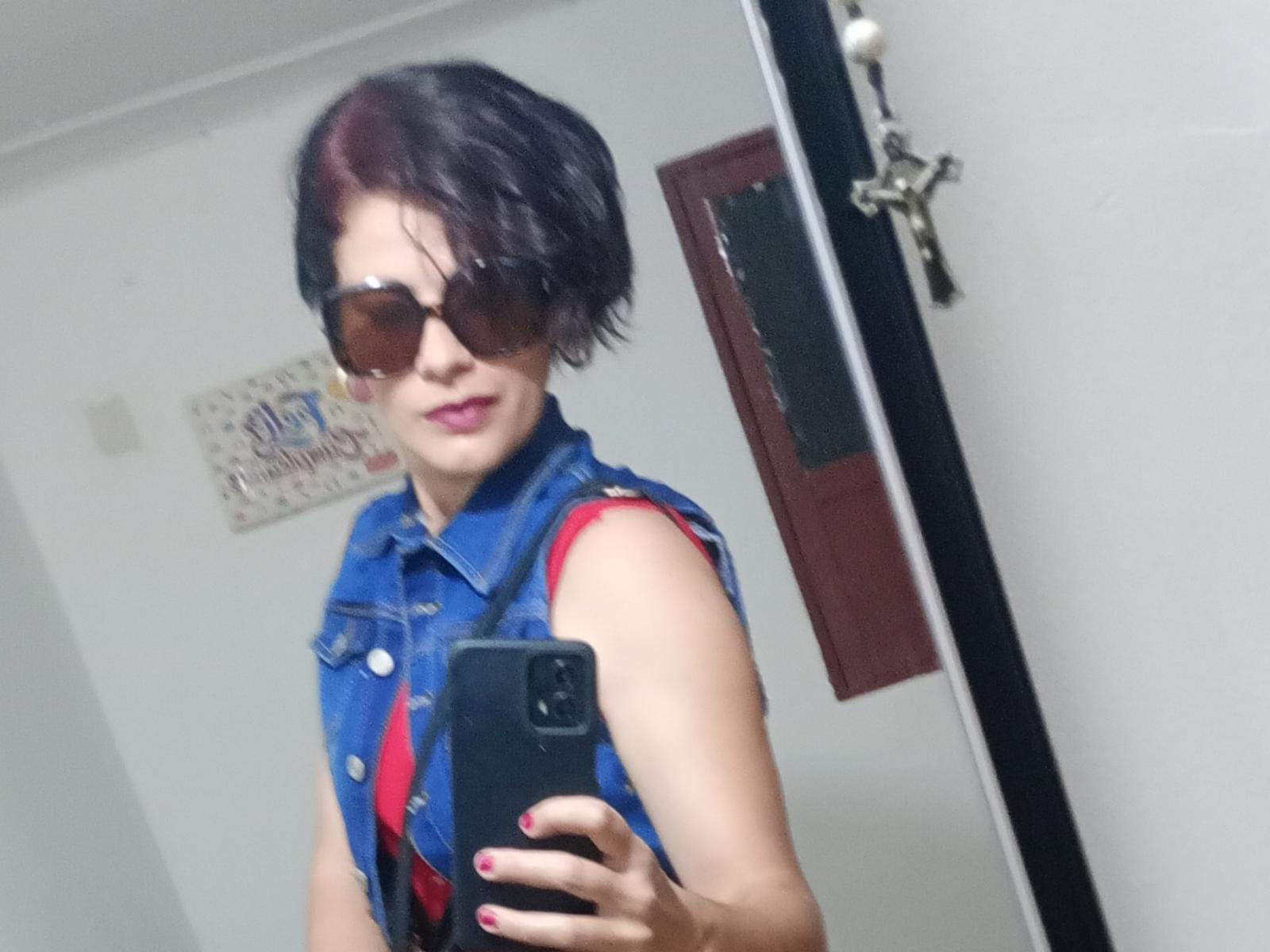LucyAndJason with short black hair, sunglasses, blue denim sleeveless shirt, red strap, holding phone, mirror selfie, pink lipstick, nail polish, white wall, door, key in background.