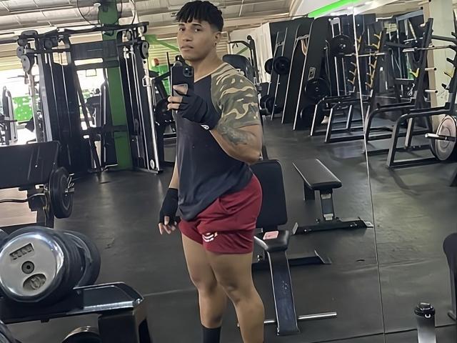 Yoximanuelprime, a young Asian male with short black hair, stands in a gym, taking a selfie with his right hand. He wears a black camo shirt, red shorts, and black wrist wraps. His left arm has a detailed tattoo. He's in the foreground, slightly to the left. The gym has various weight machines and equipment in the background. The image is a clear, well-lit photograph.