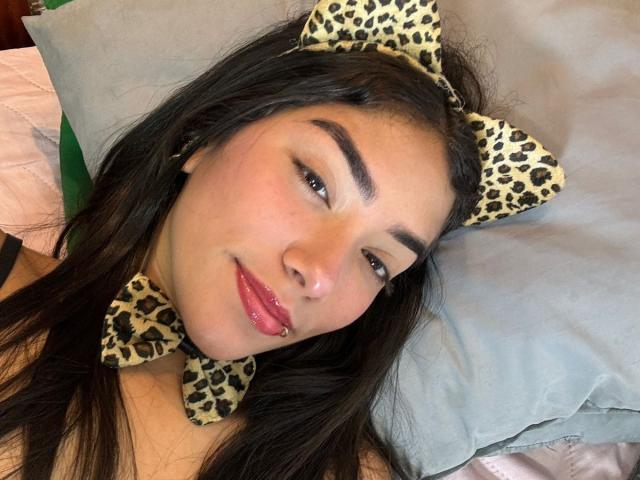 SharonKlart with leopard print cat ears and bowtie, lying on a pillow, has long black hair, pink glossy lips, and a cute smile, looking at the camera.
